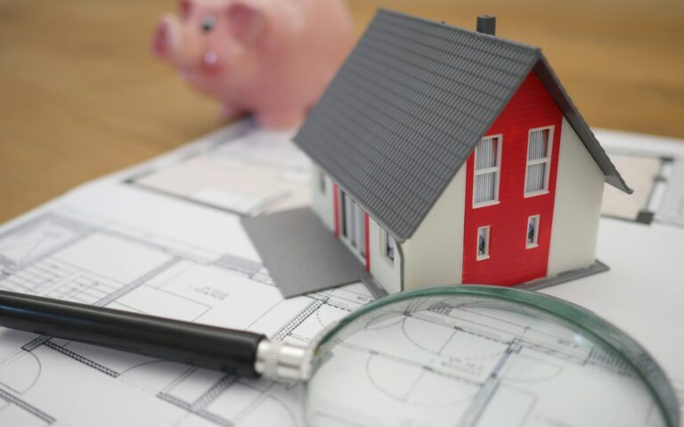 Maqueta de vivienda sobre planos con hucha y lupa